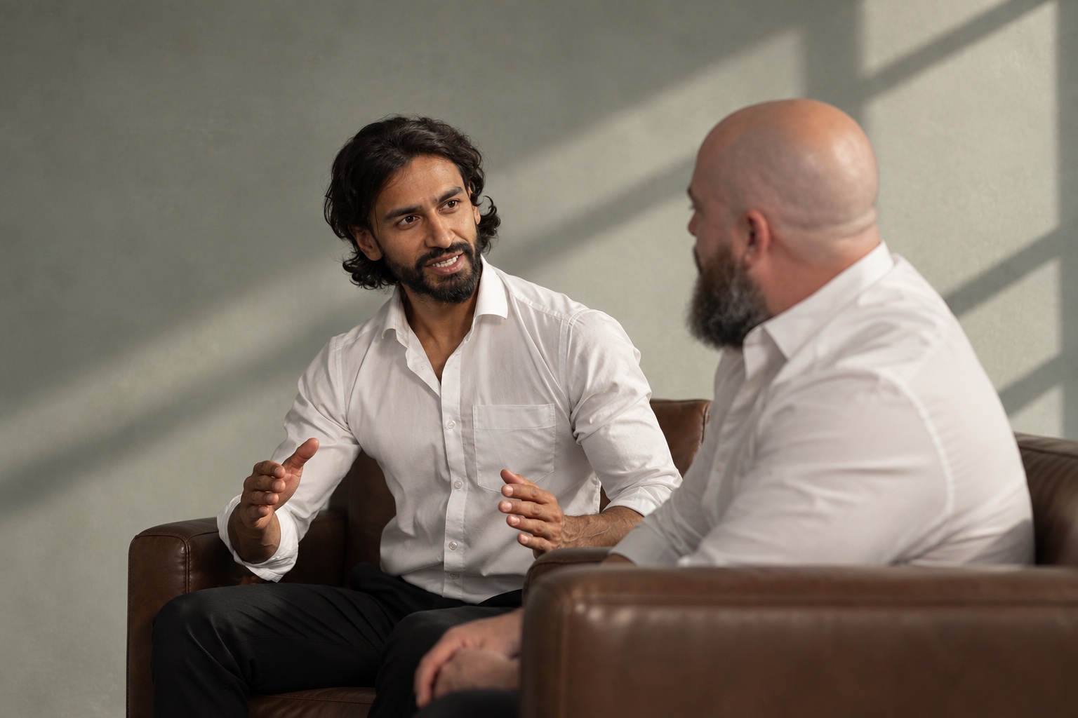 Two men in white shirts in conversation in leather armchairs, one gesturing as he talks