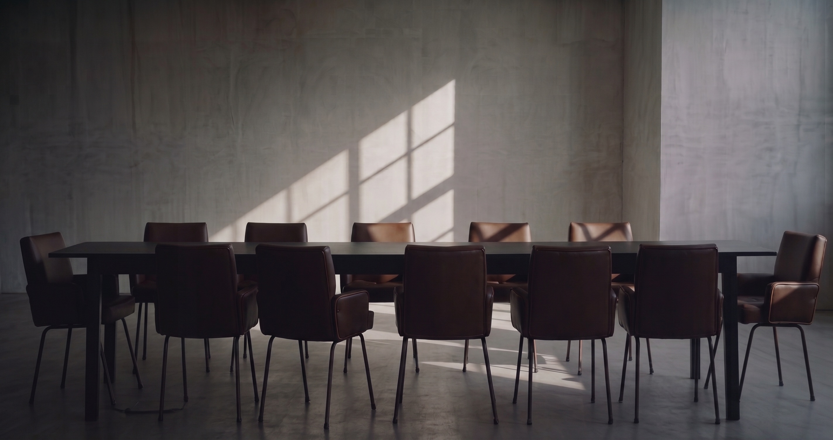 A long dark table with twelve leather chairs in a bare concrete room, sunlight falling across the far wall