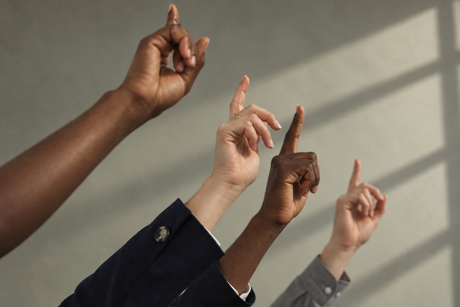 Four raised hands, index fingers up, in front of a plain wall with window light