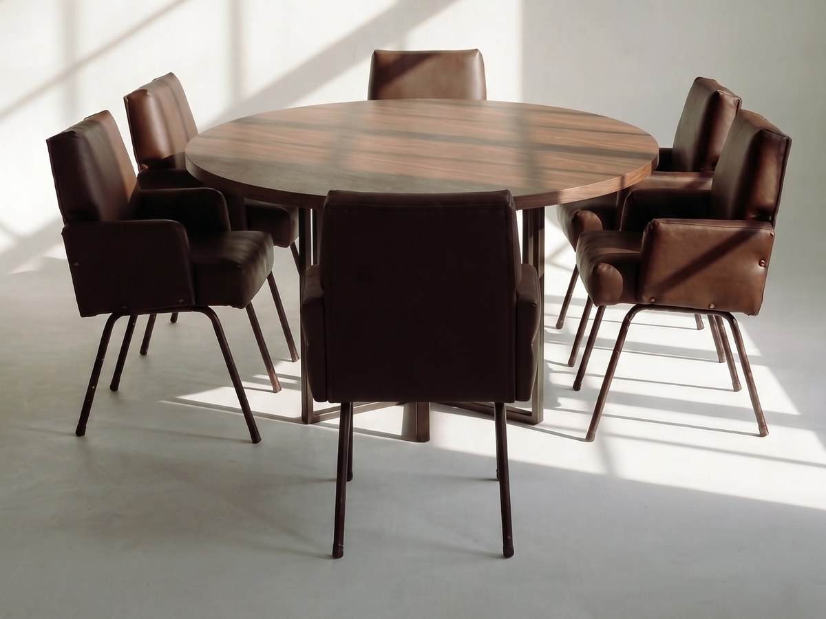 Six leather chairs around a round walnut table in a sunlit room
